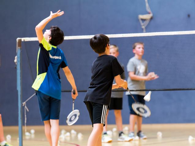 Gruppe von Kindern beim Badmintontraining in einer Sporthalle, mit Schlägern und Federbällen auf dem Boden.