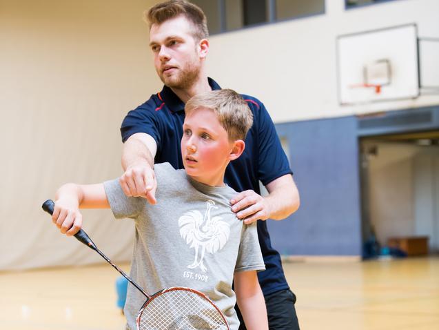 Trainingssituation: Ein Trainer zeigt einem Jungen mit Badmintonschläger die richtige Schlagtechnik in einer Sporthalle.