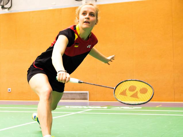 Spielerin schlägt einen Badmintonball auf einer grünen Spielfläche in einer Sporthalle mit orangefarbenen Wänden.