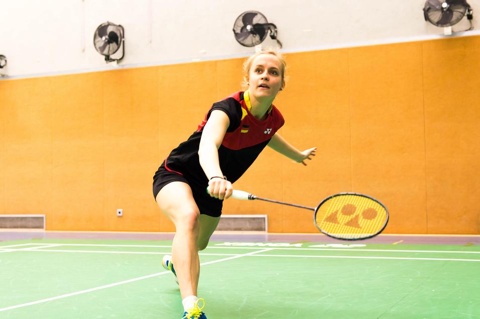 Spielerin schlägt einen Badmintonball auf einer grünen Spielfläche in einer Sporthalle mit orangefarbenen Wänden.