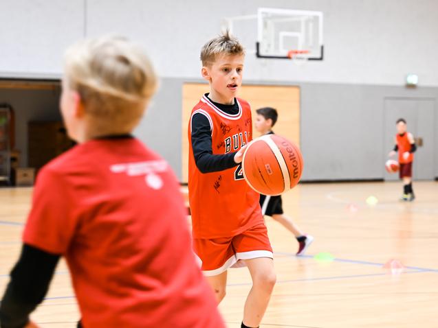 Junge in rotem Basketballtrikot dribbelt einen Ball in einer Sporthalle, während andere Kinder trainieren.