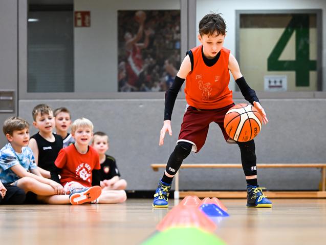 Junge übt Dribbeln mit einem Basketball, während andere Kinder am Rand zuschauen. Bunte Hütchen sind auf dem Boden angeordnet.