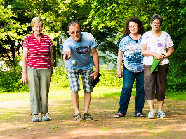 Vier Personen stehen im Freien und beobachten, wie ein Mann eine Kugel auf einen Untergrund wirft.