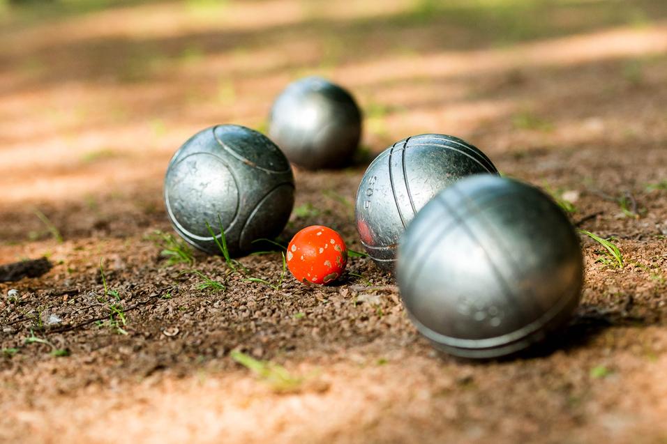 Gleise mit silbernen Boulekugeln und einer roten Kugel auf einem erdigen Boden in einem Park.