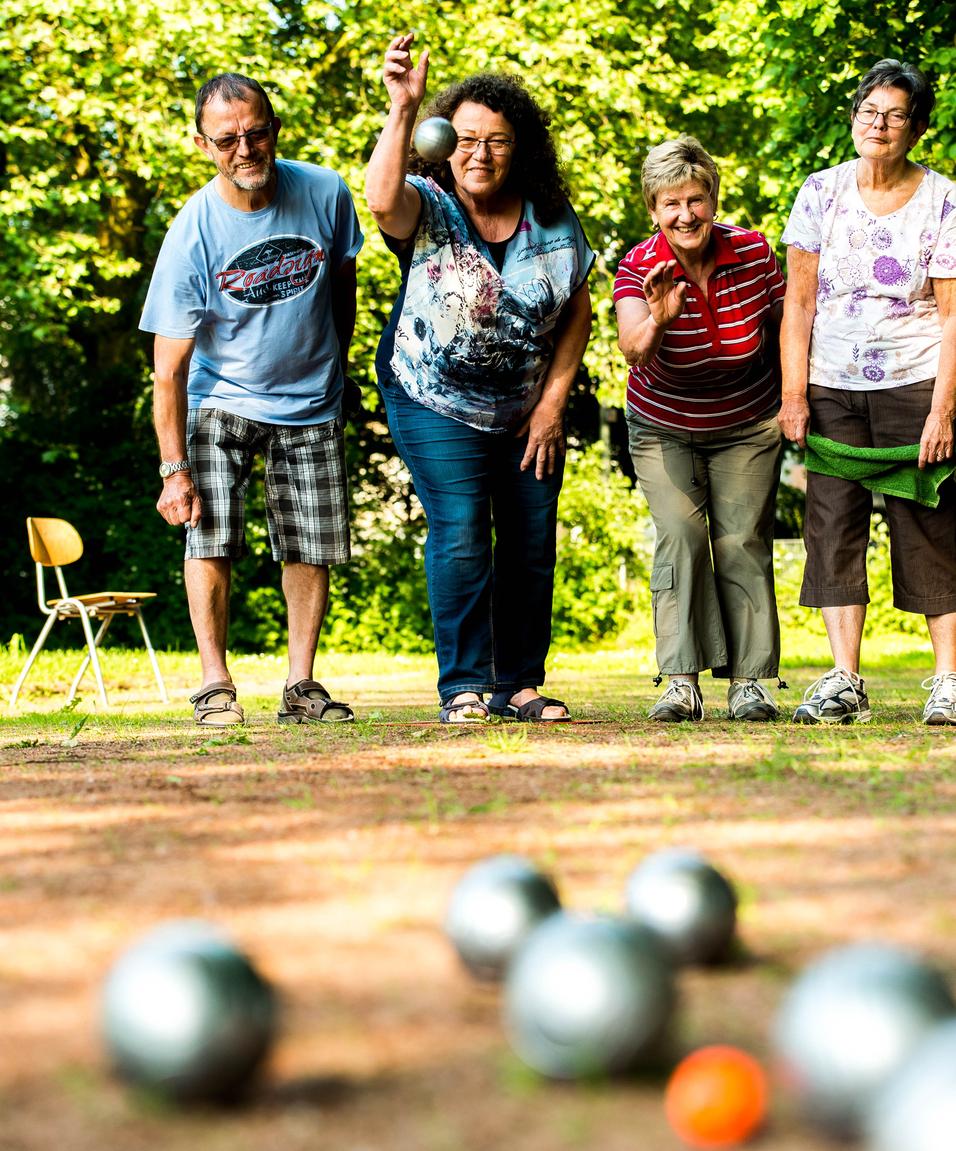 Vier Personen spielen Boule im Freien, während eine Frau gerade eine silberne Kugel wirft.