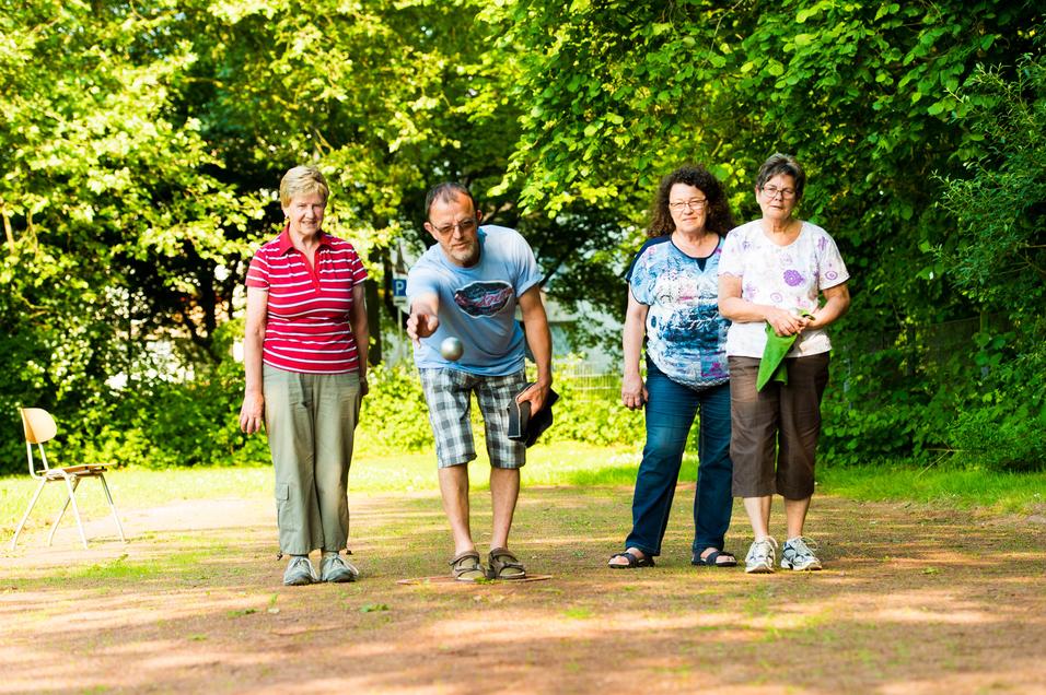 Vier Personen stehen im Freien und beobachten, wie ein Mann eine Kugel auf einen Untergrund wirft.