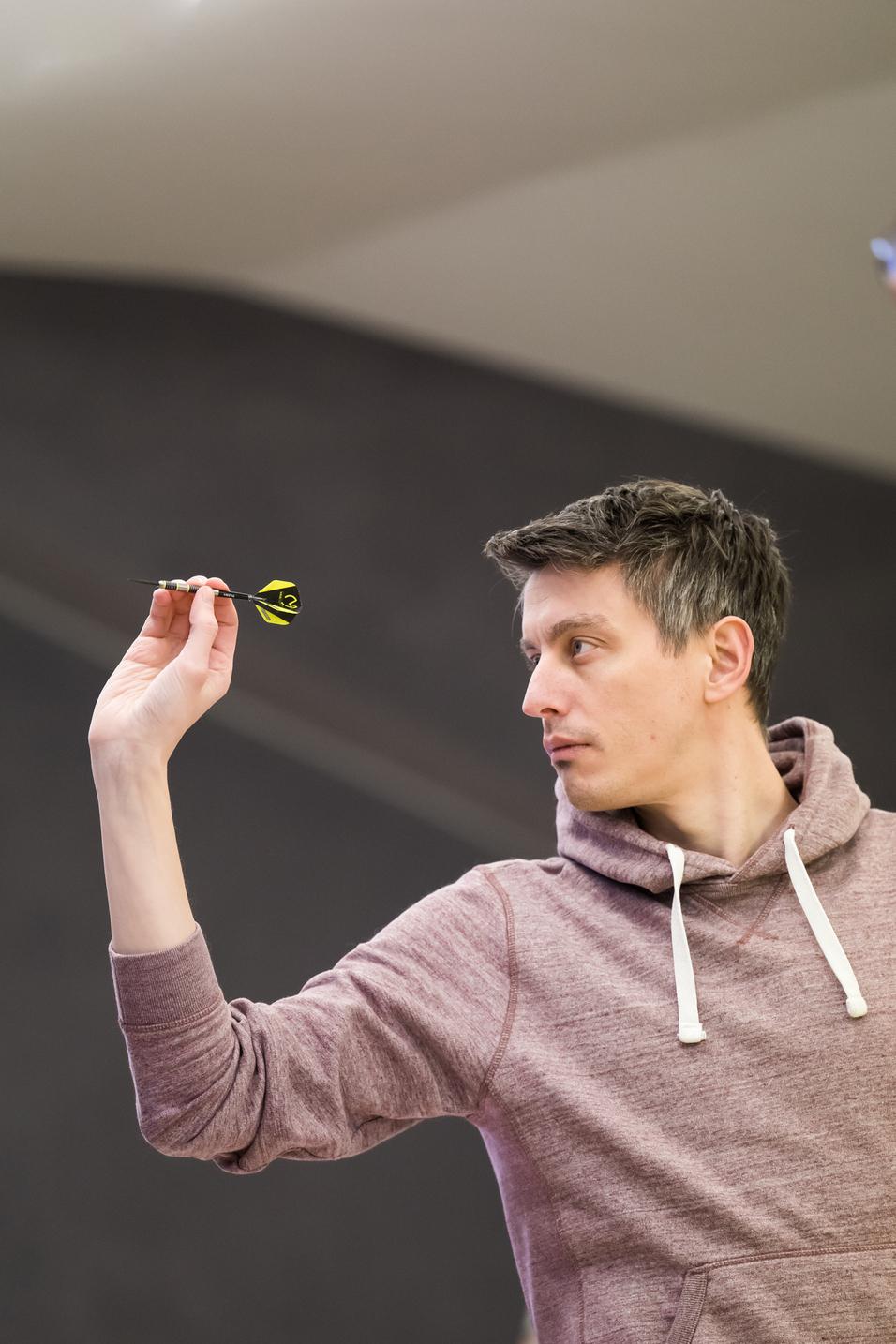 Ein Mann in einem grauen Hoodie zielt mit einem Dart auf eine Dartscheibe in einer Turnierumgebung.