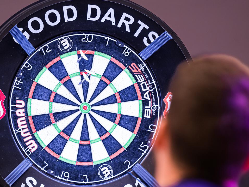 Ein Spieler wirft einen Pfeil auf ein beleuchtetes Dartboard mit dem Schriftzug „GOOD DARTS“ im Hintergrund.