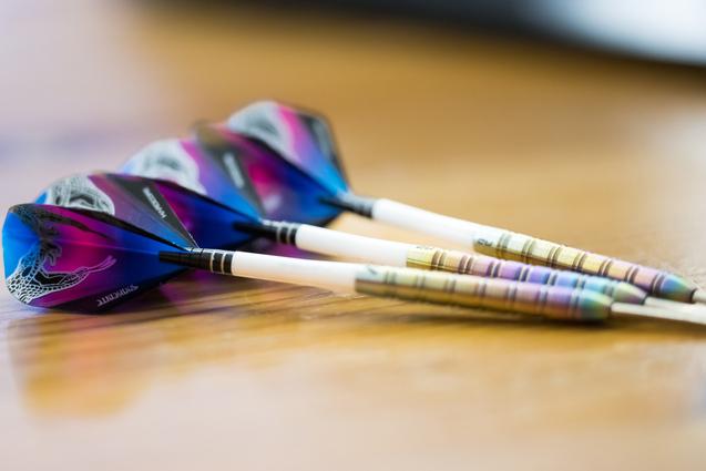 Drei Dartpfeile mit bunten Flights liegen auf einem Holztisch.