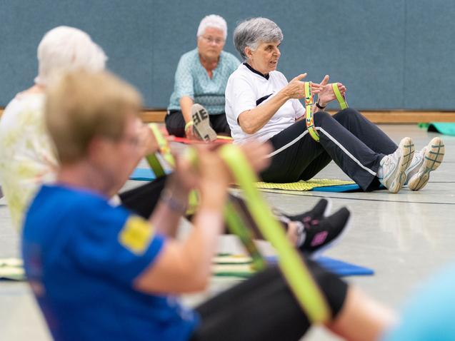 Senioren nehmen an einer Fitnessübung mit Fitnessbändern in einer Sporthalle teil, sitzend auf Fitnessmatten.