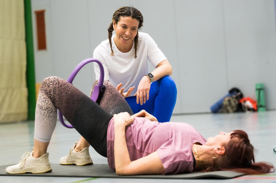 Zwei Frauen in Sportkleidung trainieren auf einer Matte, eine unterstützt die andere mit einem Fitnessgerät.