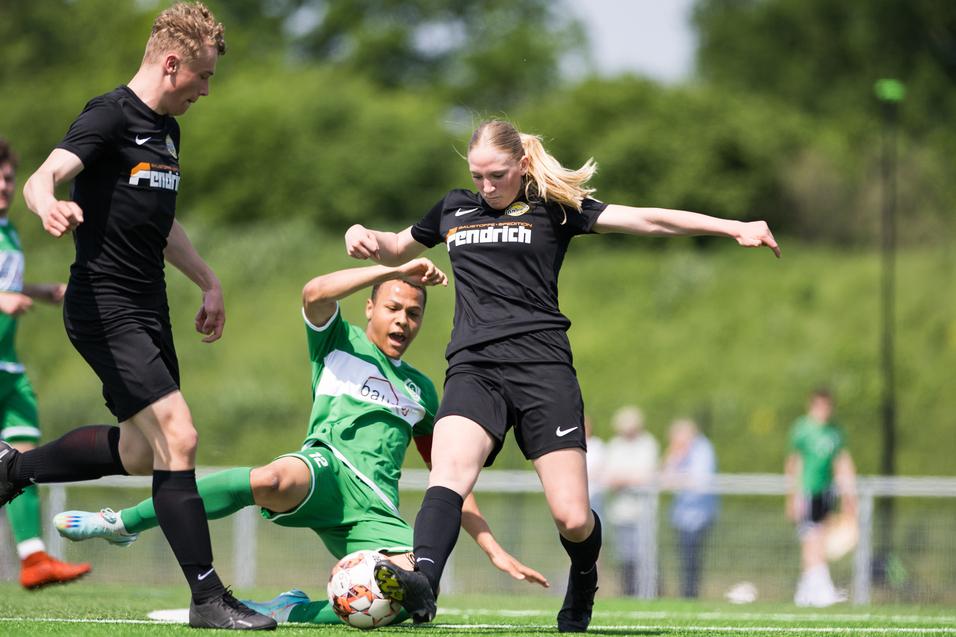 Drei Fußballspieler in Aktion auf einem Rasenplatz, eine Spielerin in Schwarz springt über einen am Boden liegenden Spieler.