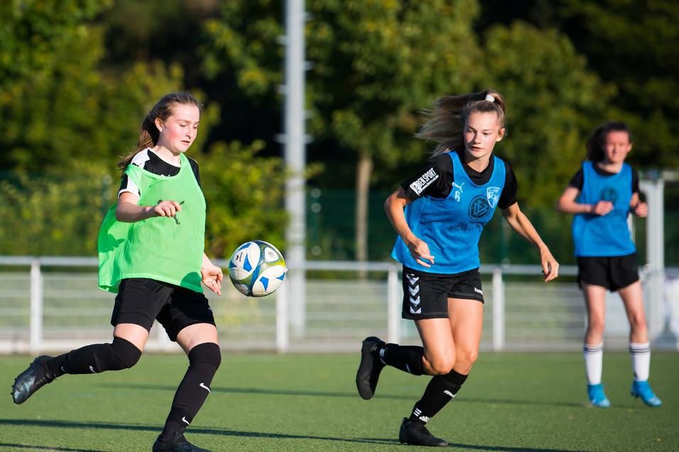 Drei Fußballspielerinnen in Trainingswesten schießen einen Ball auf einem Kunstrasenplatz unter blauem Himmel.