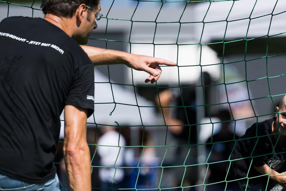 Ein Kind schaut durch ein Fußballtor, während ein Erwachsener auf es zeigt und ein Ball in der Hand hält.