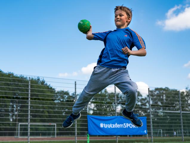 Junge springt auf einem Sportplatz und wirft einen grünen Handball, umgeben von einem Zaun und Bäumen im Hintergrund.