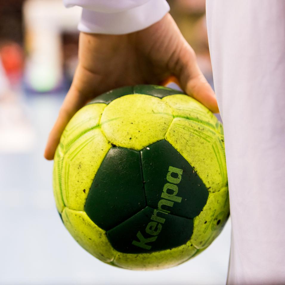 Nahaufnahme einer Hand, die einen grünen Handball mit gelben Elementen hält, im Hintergrund Spieler auf dem Spielfeld.