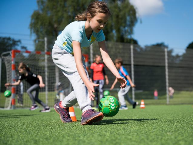 Mädchen in Sportkleidung spielt mit einem grünen Ball auf einem Kunstrasenplatz, andere Kinder trainieren im Hintergrund.