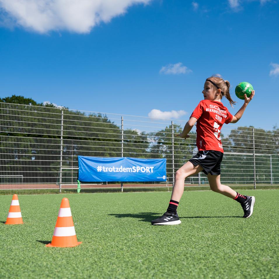 Mädchen in rotem Shirt wirft einen Handball auf einem grünen Platz, während ein Junge im Hintergrund zusieht.