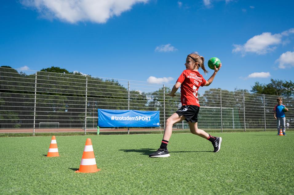 Mädchen in rotem Shirt wirft einen Handball auf einem grünen Platz, während ein Junge im Hintergrund zusieht.