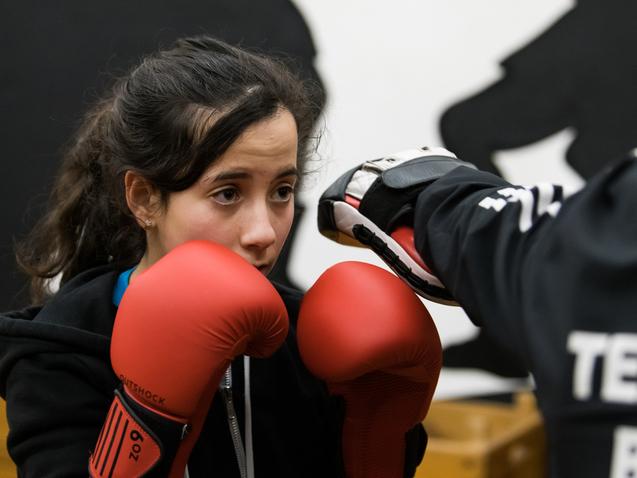 Junge Boxerin in roter Ausrüstung konzentriert auf Training mit Trainer im Hintergrund, Boxhandschuhe bereit.