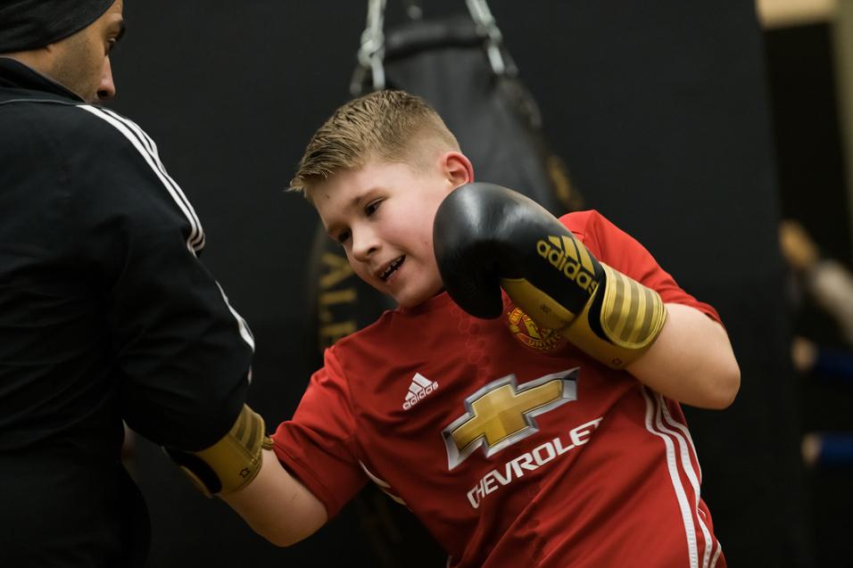 Junge in rotem Trikot mit Boxhandschuhen trainiert mit einem Trainer an einem Boxsack.
