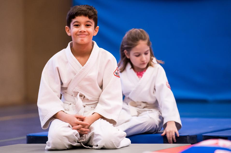 Zwei Kinder in weißen Judoanzügen sitzen auf einer Matte in einem Judoraum, der Hintergrund ist blau.