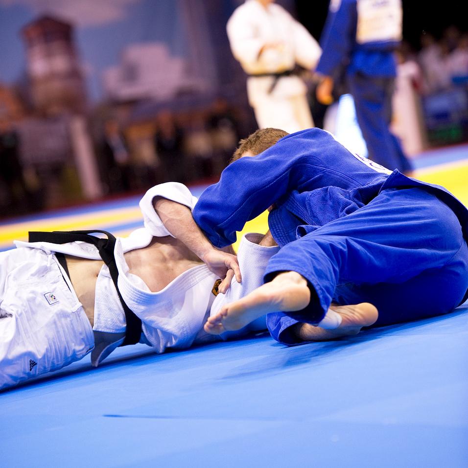 Zwei Judoka kämpfen auf einer blauen Matte, einer in weißem und der andere in blauem Gi, während Zuschauer im Hintergrund zuschauen.