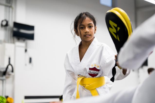 Junge Karatekämpferin in weißem Gi mit gelben Gürteln übt den Kick gegen ein Schlagpolster in einer Trainingshalle.