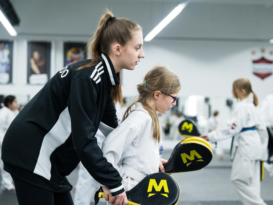 Trainerin unterstützt ein Kind mit einer Schlagpuppe in einer Kampfsport-Trainingsumgebung.