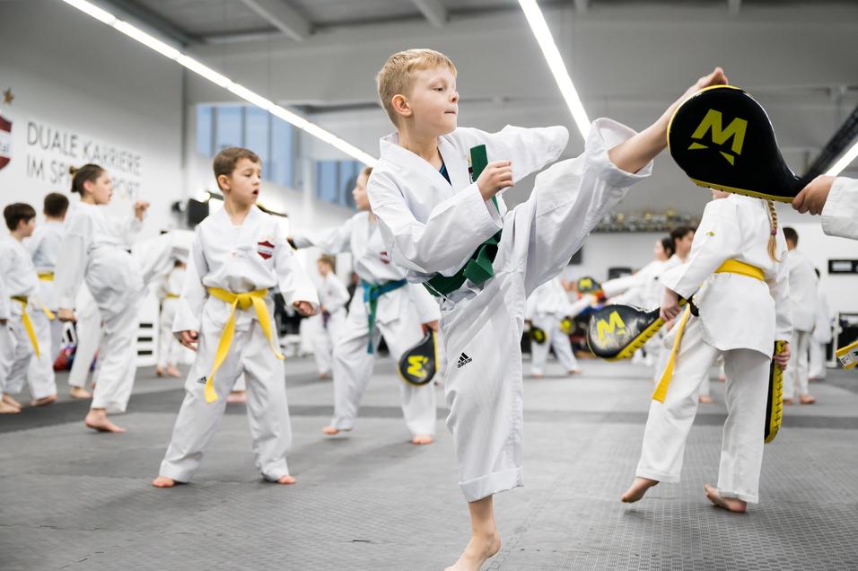 Eine Gruppe von Kindern in Karate-Uniformen übt in einem Trainingsraum, ein Junge führt einen Kick aus.