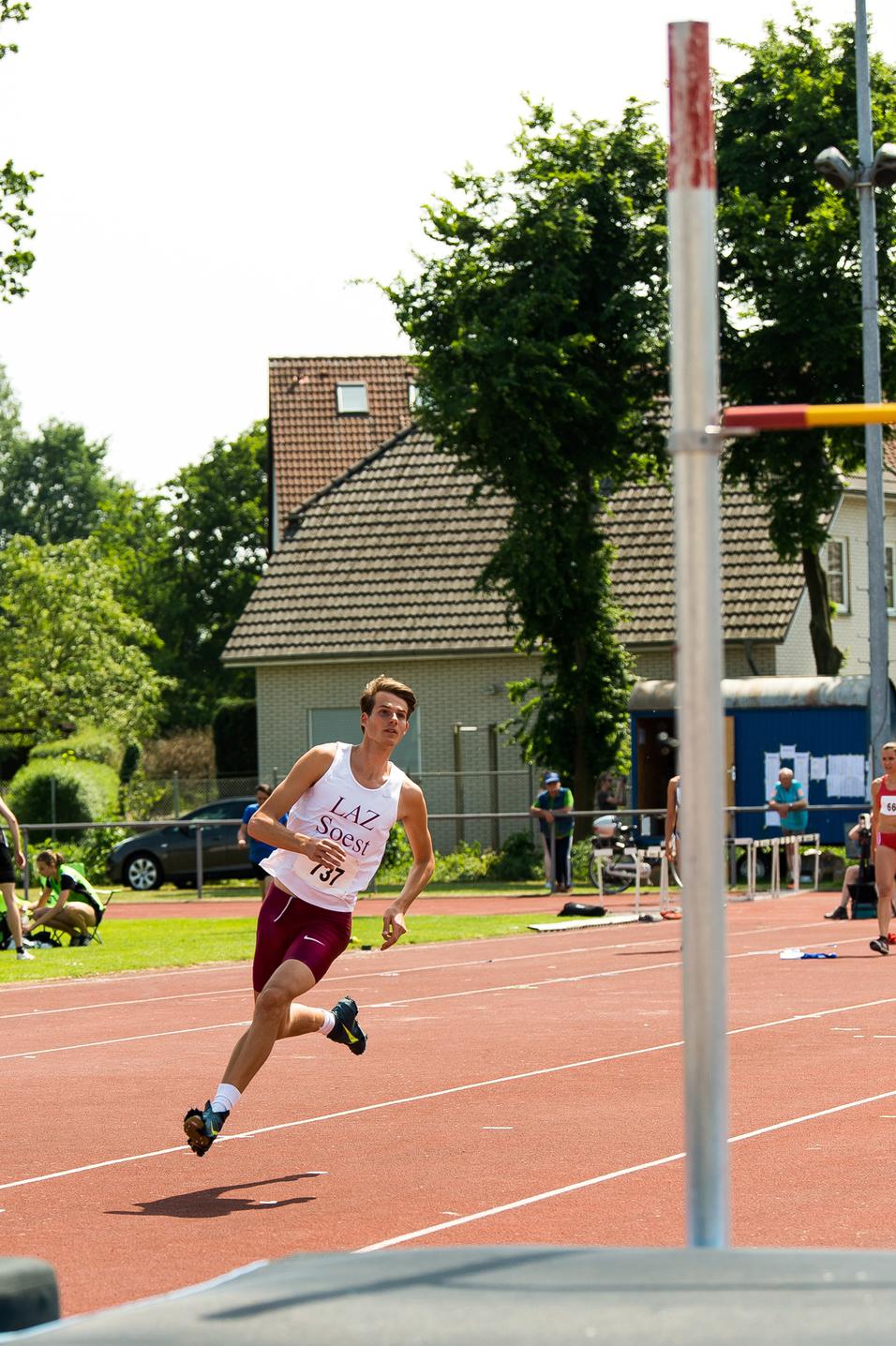 Athlet in Sportkleidung sprintet auf einer Leichtathletikbahn, im Hintergrund Zuschauer und eine Hochsprung-Anlage.