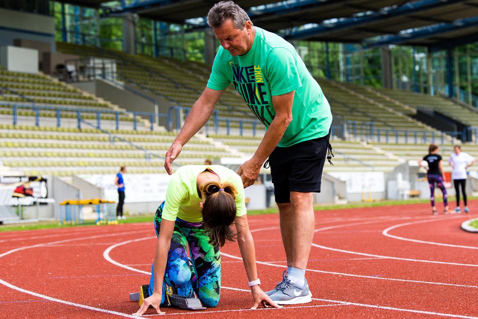 Trainer hilft Sportlerin beim Startblock auf einer Laufbahn in einem Stadion. Im Hintergrund stehen weitere Athleten.