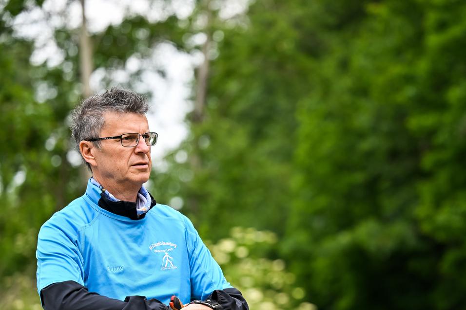 Mann in blauer Sportjacke hält Nordic Walking-Stöcke in einer grünen, bewaldeten Umgebung.
