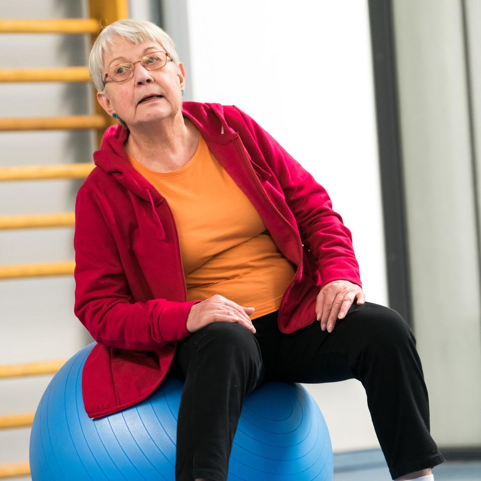 Ältere Frau in roter Jacke sitzt auf einem blauen Gymnastikball in einer Sporthalle.