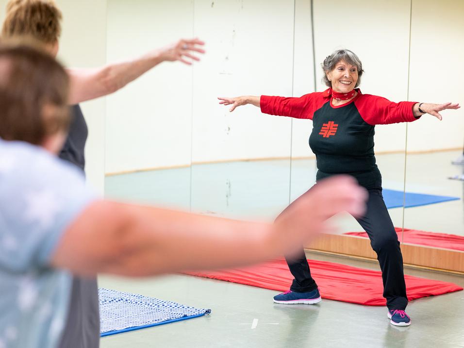 Eine ältere Frau in Sportkleidung führt eine Dehnübung in einem Fitnessraum mit Spiegeln aus.