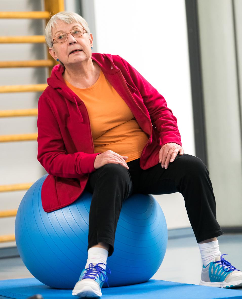 Ältere Frau in roter Jacke sitzt auf einem blauen Gymnastikball in einer Sporthalle.