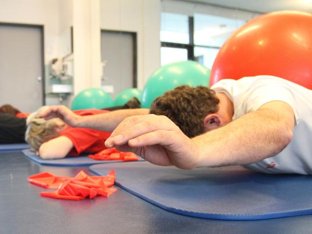 Gruppentraining mit Personen liegend auf Matten, während Bälle im Hintergrund sichtbar sind.