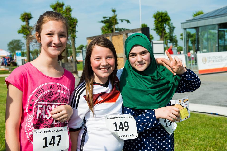 Drei lächelnde Mädchen in Sportkleidung mit Startnummern stehen auf einer Wiese, umgeben von einer Veranstaltung.
