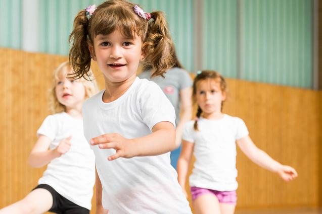 Vier Kinder in Sportkleidung bewegen sich in einer Turnhalle, ein Mädchen mit Zöpfen lächelt in die Kamera.