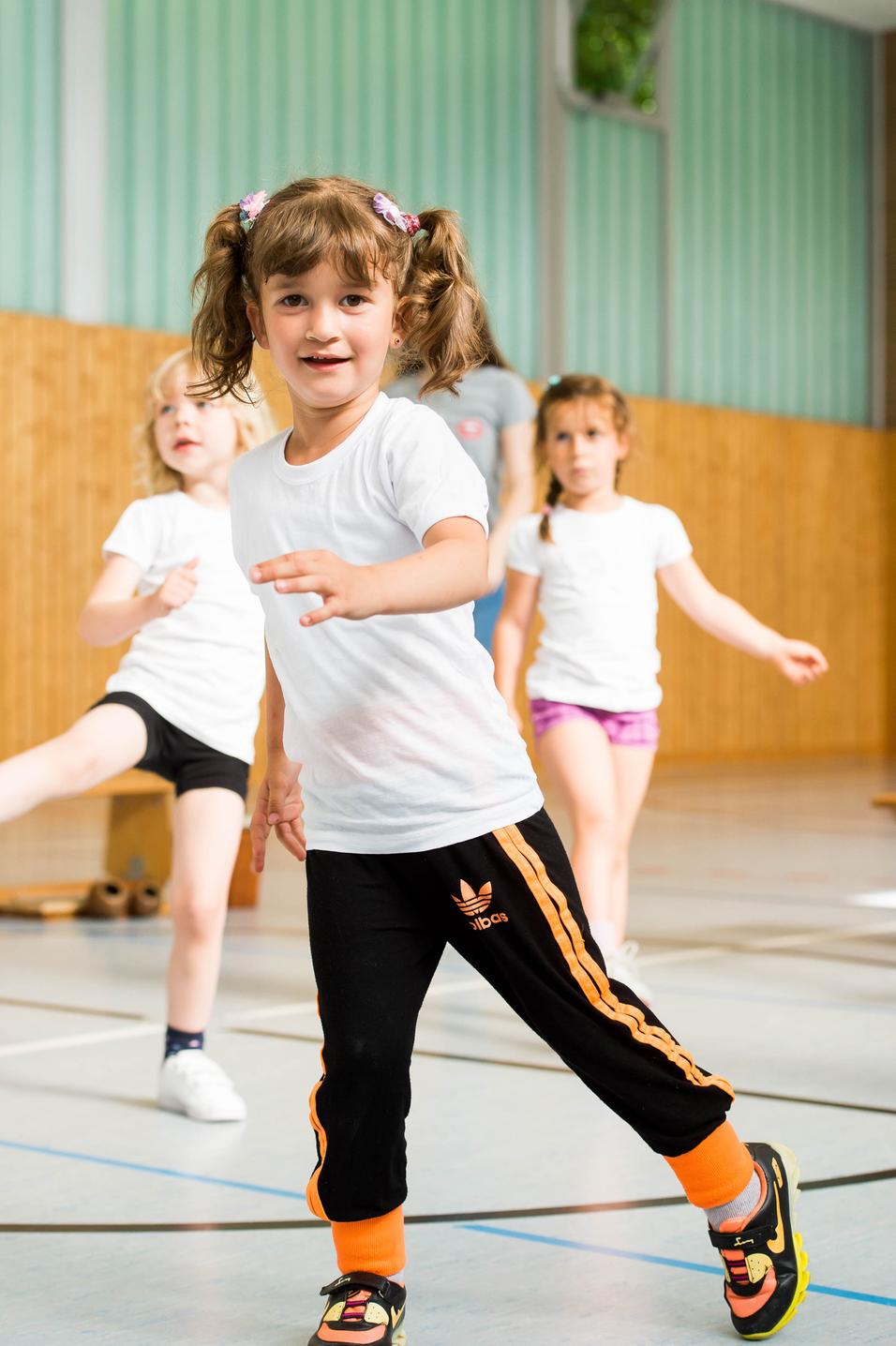 Vier Kinder in Sportkleidung bewegen sich in einer Turnhalle, ein Mädchen mit Zöpfen lächelt in die Kamera.