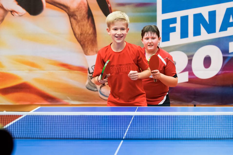 Zwei Kinder in roten Trikots stehen lächelnd an einem Tischtennistisch mit Schlägern in den Händen.