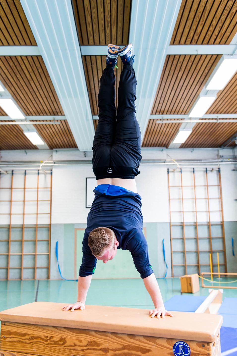 Sportler führt einen Handstand auf einer Kiste in einer Turnhalle durch, im Hintergrund sind Turngeräte sichtbar.