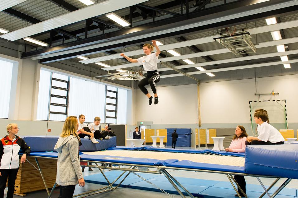 Junge springt auf einem Trampolin, während Zuschauer und Trainer im Hintergrund beobachten. Sporthalle mit hellem Licht.