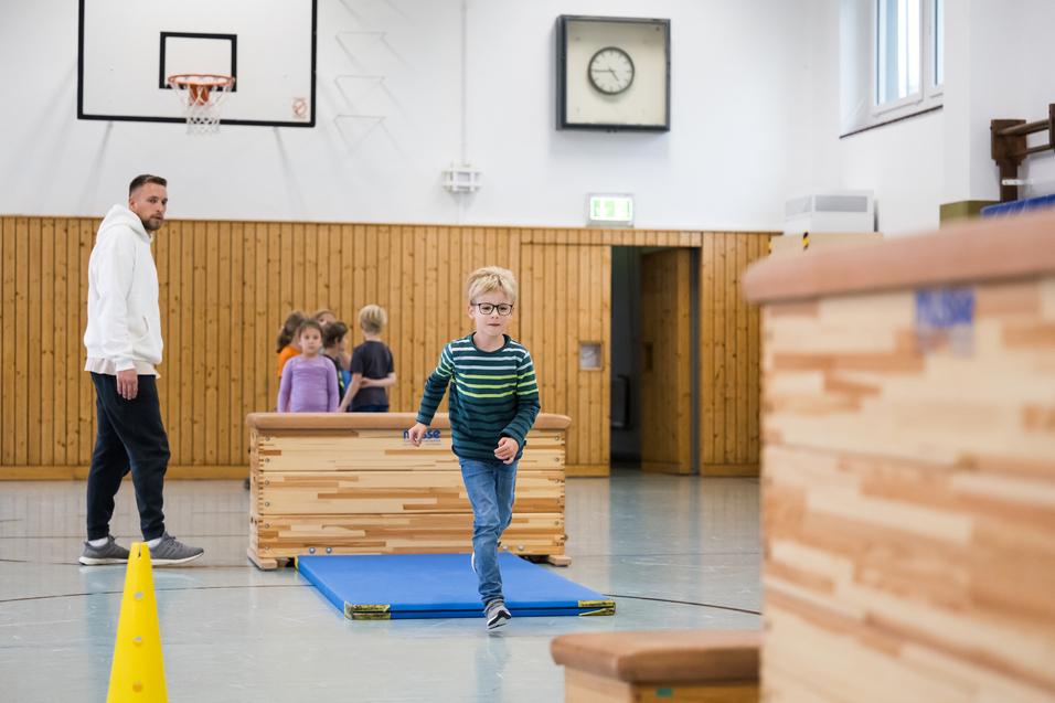 Ein Junge in gestreiftem Pullover läuft über eine Übungsfläche, während Erwachsene und Kinder im Hintergrund aktiv sind.