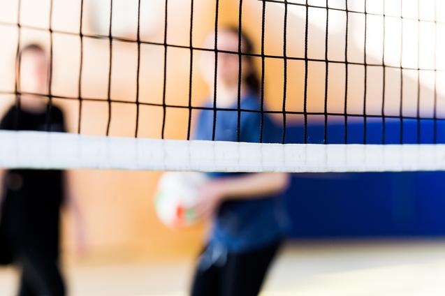 Unscharfe Silhouetten von zwei Personen hinter einem Volleyballnetz in einer Sporthalle. Eine Person hält einen Volleyball.