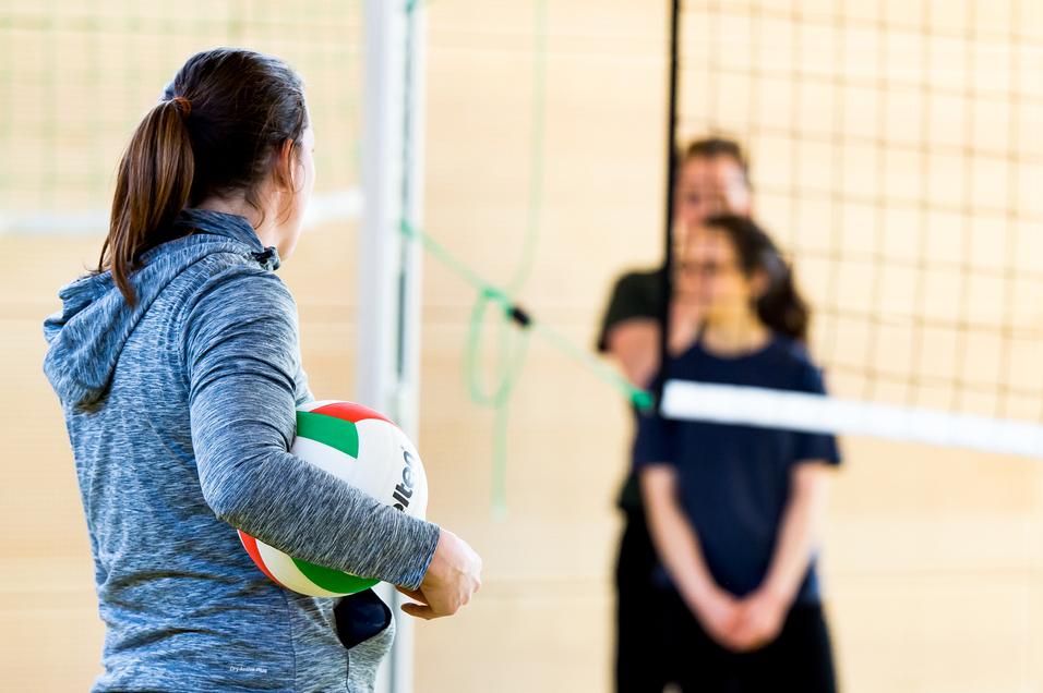 Eine Person mit einem Volleyball steht vor dem Netz, während zwei weitere Personen im Hintergrund warten.