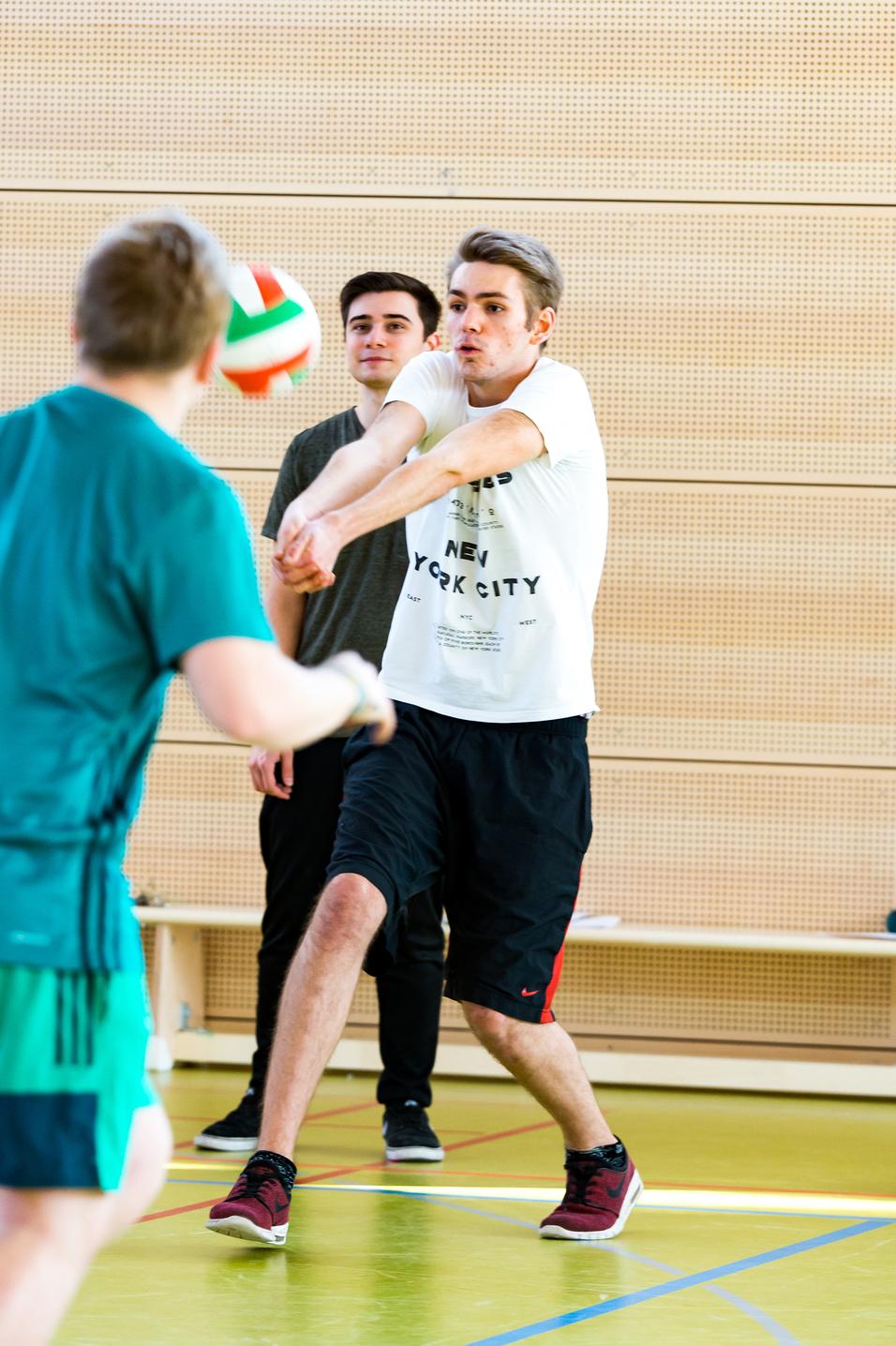 Junger Mann in Sportbekleidung spielt Volleyball in einer Turnhalle, während ein weiterer Spieler im Hintergrund zuschaut.