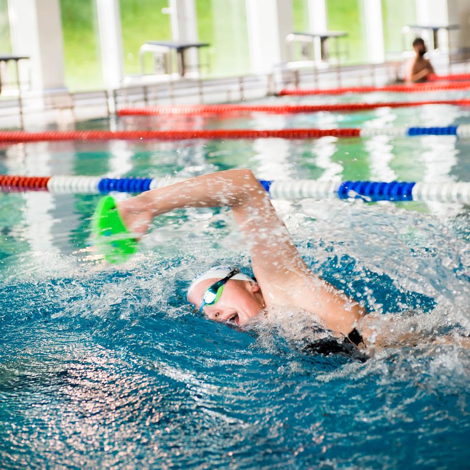 Schwimmerin mit grüner Paddel-Handschuh im Wasser eines Hallenbads, umgeben von bunten Bahnen und Fenstern.