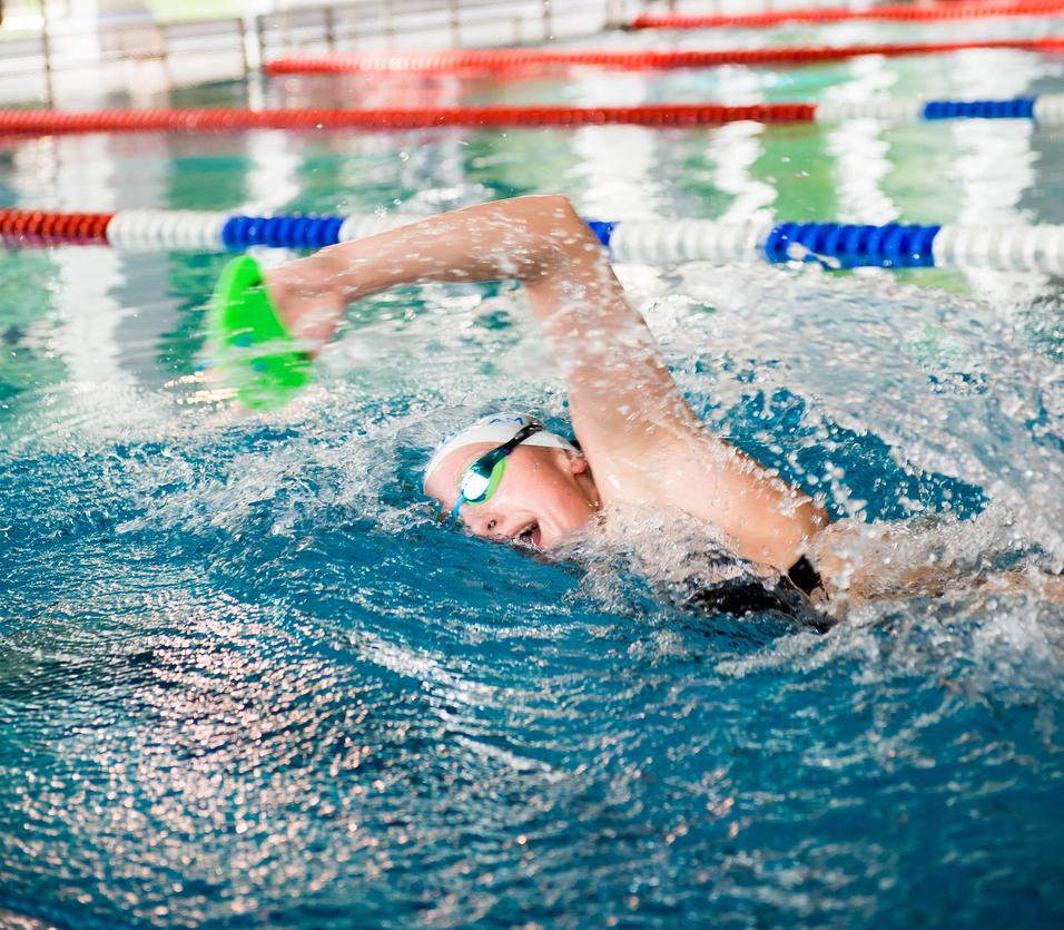Schwimmerin mit grüner Paddel-Handschuh im Wasser eines Hallenbads, umgeben von bunten Bahnen und Fenstern.