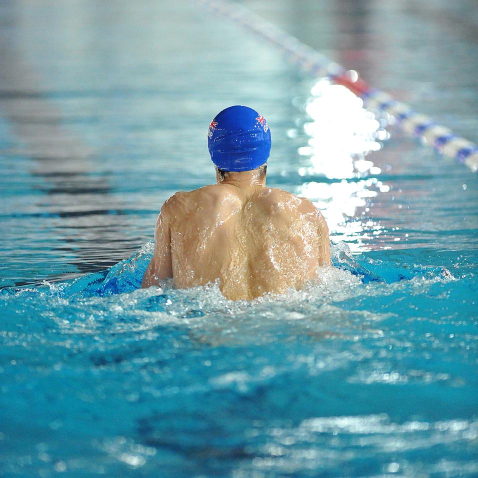 Schwimmer mit blauer Badehaube gleitet im Wasser eines Schwimmbeckens, Rücken zur Kamera.
